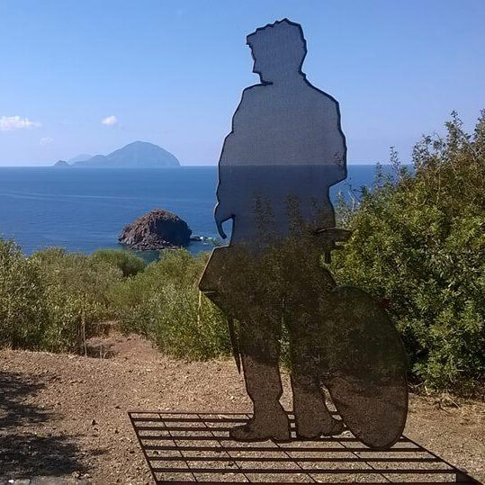 L'Ombra del Postino: a steel silhouette of a postman beside his bicycle wheel, installed on a headland above the sea with the Aeolian islands behind.