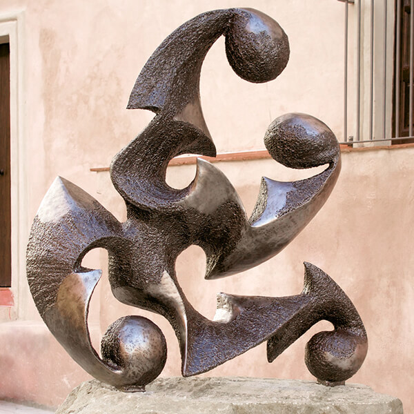 Mater la Pietà: an abstract iron sculpture of interlocking curved blades and burnished spheres, standing on a stone plinth against a pink wall.