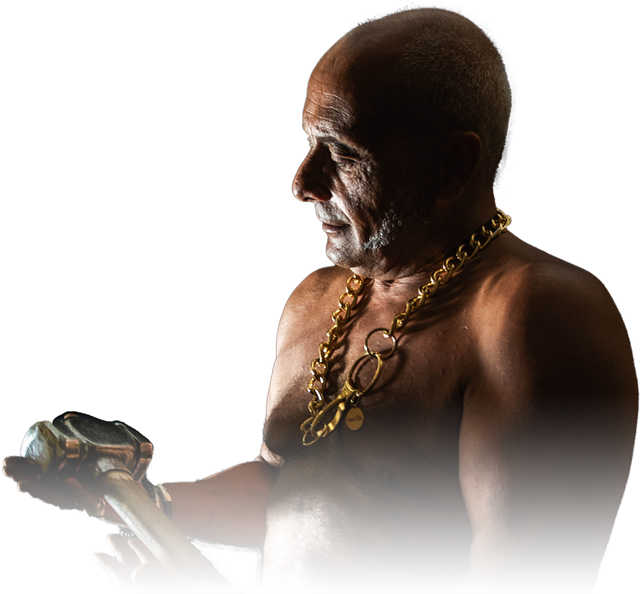 Fabio Pilato in profile in a darkened room, bare-chested and wearing a heavy gold chain, turning a blacksmith&rsquo;s hammer over in his hands.