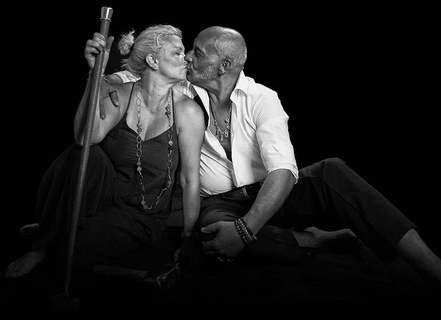 Black and white portrait: Fabio Pilato leans in to kiss a woman seated beside him, who holds a long-handled hammer upright.