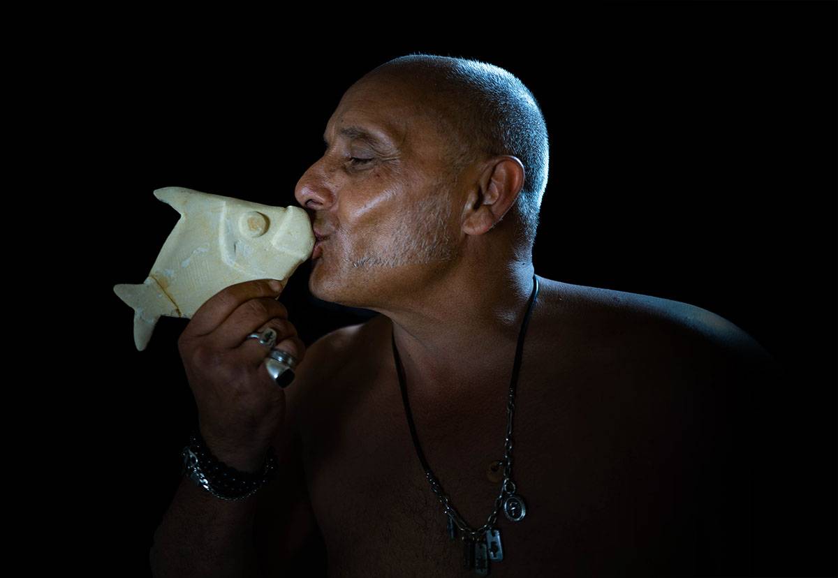 Fabio Pilato in profile, kissing the snout of a small pale fish sculpture he holds up to his mouth.